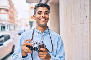 Young latin tourist man smiling happy using vintage camera walking at the city.