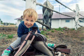 Cute adorable caucasian little smiling toddler portrait boy sitting on saddle on ground enjoy having fun riding supposed horse on countryside farm outdoors. Happy childhood on country ranch