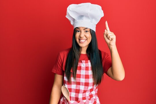 Young beautiful hispanic girl wearing baker uniform and cook hat pointing finger up with successful idea. exited and happy. number one.