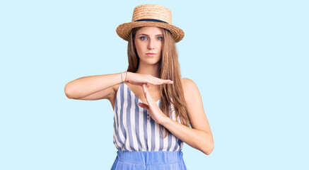 Obraz na płótnie Canvas Young beautiful blonde woman wearing summer hat doing time out gesture with hands, frustrated and serious face