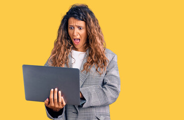 Young hispanic woman with tattoo holding laptop scared and amazed with open mouth for surprise, disbelief face