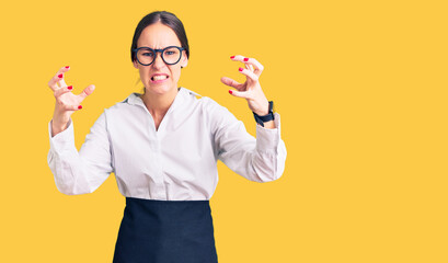 Beautiful brunette young woman wearing professional waitress apron shouting frustrated with rage, hands trying to strangle, yelling mad