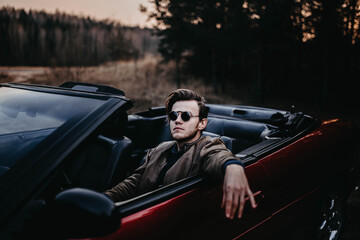 brutal man with a beard sits in a red convertible in sunglasses in nature. Male bachelor and...
