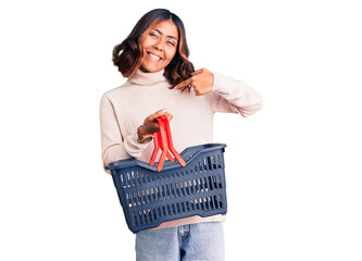 Young beautiful mixed race woman holding supermarket shopping basket pointing finger to one self smiling happy and proud