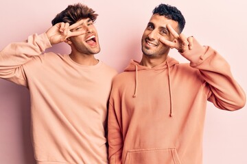 Young gay couple wearing casual clothes doing peace symbol with fingers over face, smiling cheerful showing victory