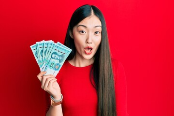 Young chinese woman holding 50 yuan chinese banknotes scared and amazed with open mouth for surprise, disbelief face