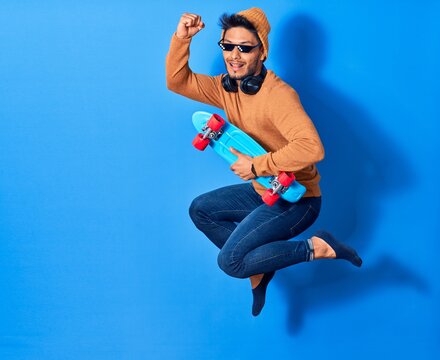 Young handsome latin man wearing wool cap and thug life glasses.  Jumping with smile on face holding skate over isolated blue background