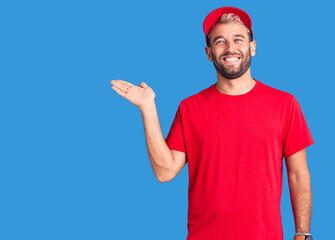 Naklejka premium Young handsome blond man wearing t-shirt and cap smiling cheerful presenting and pointing with palm of hand looking at the camera.