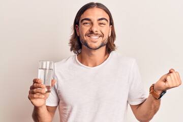 Young handsome man drinking glass of water screaming proud, celebrating victory and success very excited with raised arm