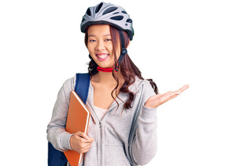 Young beautiful chinese girl wearing student backpack and bike helmet holding book celebrating victory with happy smile and winner expression with raised hands