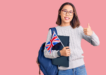 Young beautiful chinese girl wearing student backpack holding binder and uk flag smiling happy and positive, thumb up doing excellent and approval sign