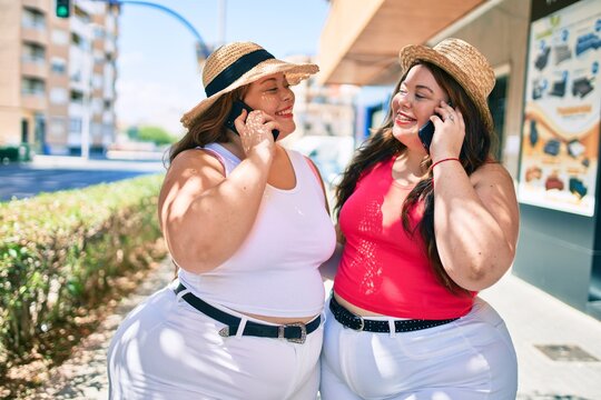 Two plus size overweight sisters twins women speaking on the phone outdoors on a sunny day