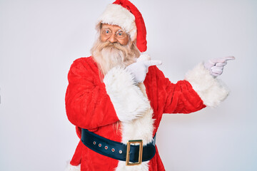 Old senior man with grey hair and long beard wearing santa claus costume smiling and looking at the camera pointing with two hands and fingers to the side.