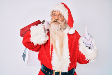 Old senior man with grey hair and long beard wearing santa claus costume holding shopping bags surprised with an idea or question pointing finger with happy face, number one