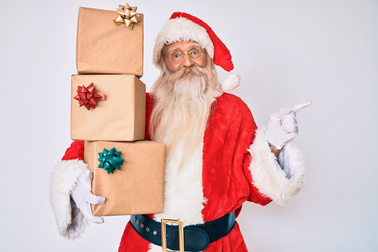 Old senior man with grey hair and long beard wearing santa claus costume holding presents smiling happy pointing with hand and finger to the side