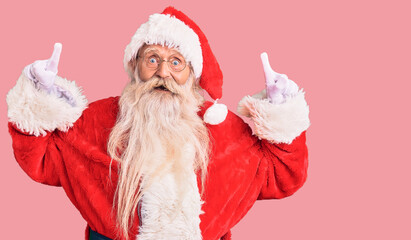 Old senior man with grey hair and long beard wearing traditional santa claus costume smiling amazed and surprised and pointing up with fingers and raised arms.