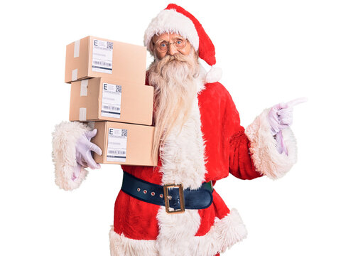 Old senior man with grey hair and long beard wearing santa claus costume holding boxes smiling happy pointing with hand and finger to the side