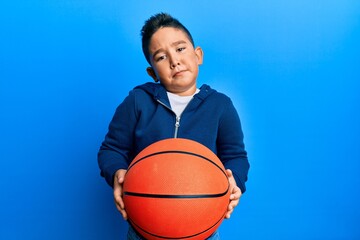 Little boy hispanic kid holding basketball ball clueless and confused expression. doubt concept.