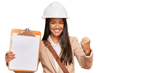 Beautiful hispanic woman architect showing black space on clipboard screaming proud, celebrating victory and success very excited with raised arms