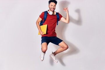 Young handsome hispanic student man wearing backpack smiling happy. Jumping with smile on face holding book using headphones. Celebrating with fist up over isolated white background.