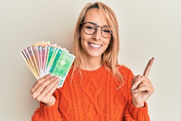 Beautiful blonde woman holding norwegian krone banknotes smiling happy pointing with hand and finger to the side