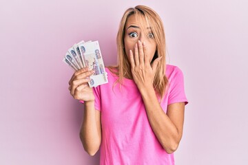 Beautiful blonde woman holding 500 russian ruble banknotes covering mouth with hand, shocked and afraid for mistake. surprised expression