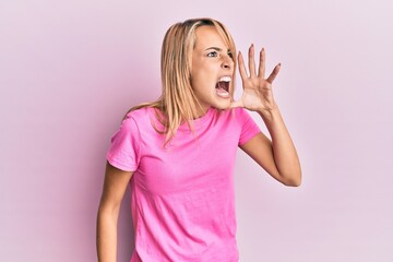 Beautiful blonde woman wearing casual pink tshirt shouting and screaming loud to side with hand on mouth. communication concept.