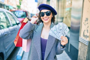 Young beautiful girl smiling happy with french style holding shopping bags and dollars banknotes at street of city