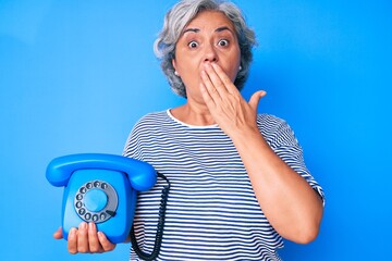 Senior hispanic grey- haired woman holding vintage telephone covering mouth with hand, shocked and afraid for mistake. surprised expression