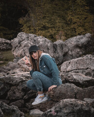 Naklejka na ściany i meble Attractive brunette sitting on a rock in the nature, wearing a blue jeans outfit, making a peace gesture as a hello as she spots the camera looking at her