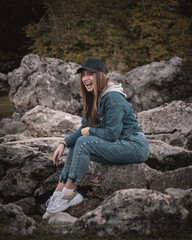 Attractive brunette wearing a jeans outfit and a black cap, sitting on a rock in the nature looking at something, caught in the moment of genuine pure beautiful laughter, making a funny expression