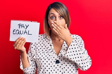 Young beautiful woman holding pay equal message paper covering mouth with hand, shocked and afraid for mistake. surprised expression