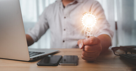 Male hand holding a light bulb And there is a gear icon in it And he is using a notebook computer....