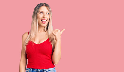 Young beautiful blonde woman wearing casual clothes smiling with happy face looking and pointing to the side with thumb up.