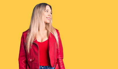 Young beautiful blonde woman wearing red leather jacket looking away to side with smile on face, natural expression. laughing confident.