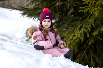 Fototapeta na wymiar Cute little girl sits in the snow near green fir trees. Ski resort. A child in overalls against the background of a winter landscape. Winter walks, fun outdoors winter time.