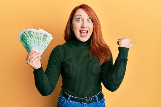 Beautiful redhead woman holding 5 singapore dollars banknotes pointing thumb up to the side smiling happy with open mouth