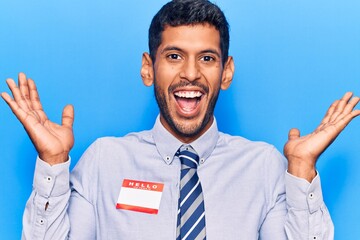 Young latin man wearing sticker with hello my name is message celebrating victory with happy smile and winner expression with raised hands