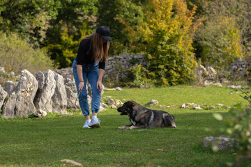 Naklejka na ściany i meble Brunette girl playing with a wild dog in a field surrounded by rocks. Holding a stick that the dog is trying to catch. Playing a typical game of fetch with a energetic dog
