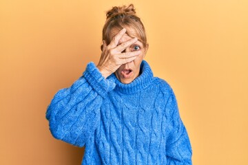 Middle age blonde woman wearing casual winter sweater peeking in shock covering face and eyes with hand, looking through fingers afraid