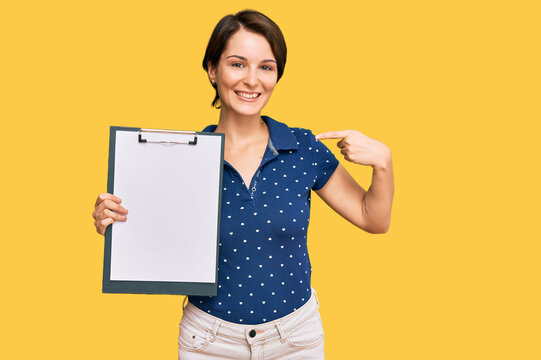 Young brunette woman with short hair holding clipboard with blank space pointing finger to one self smiling happy and proud