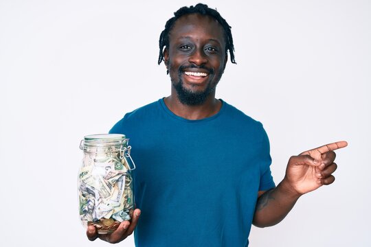Young african american man with braids holding jar with savings smiling happy pointing with hand and finger to the side