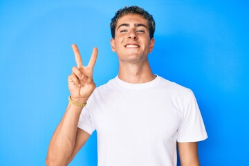 Young handsome man wearing casual white t shirt smiling with happy face winking at the camera doing victory sign. number two.