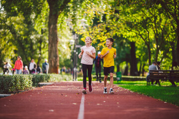 Couple of kids boy and girl doing cardio workout, jogging in park on jogging track red. Cute twins runing together. Run children, young athletes. Teen brother and sister running along path outdoors
