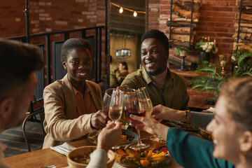Portrait of young African-American couple clinking glasses while enjoying dinner party with friends and family in cozy interior
