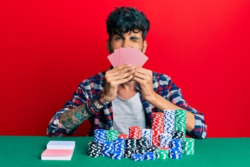 Young hispanic man playing gambling poker covering face with cards winking looking at the camera...