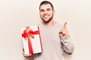 Young handsome man holding gift smiling with an idea or question pointing finger with happy face, number one