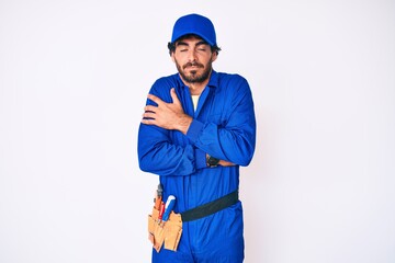 Handsome young man with curly hair and bear weaing handyman uniform shaking and freezing for winter cold with sad and shock expression on face