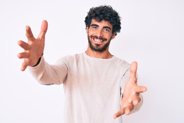 Handsome young man with curly hair and bear wearing casual winter sweater looking at the camera smiling with open arms for hug. cheerful expression embracing happiness.