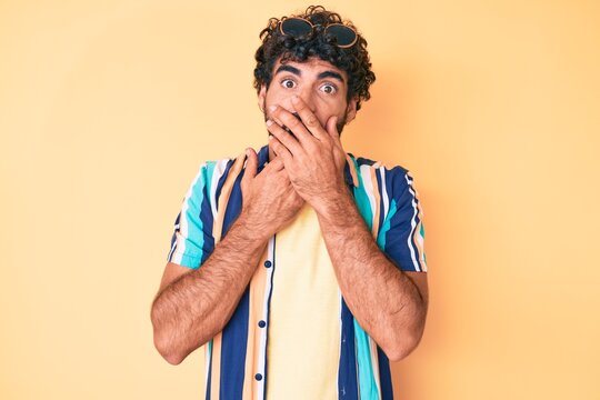 Handsome young man with curly hair and bear wearing summer shirt and sunglasses shocked covering mouth with hands for mistake. secret concept.